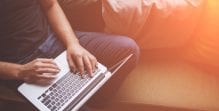 A man sits on a sofa with his hands typing on a laptop