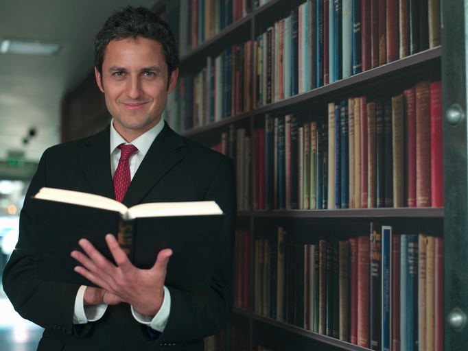 A man standing in front of a bookcase full of books holding an open book