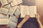 A man is sat on the floor and is reading an open book on his lap with other books surrounding him on the floor