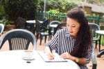 A young woman writing a law firm application outdoors with a coffee on the table.