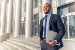 A man is standing on some steps outside of his office building dressed in a suit and holding a laptop or notebook