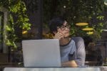 A man is sat a a table on his laptop and looking to the right with a leafy background behind him