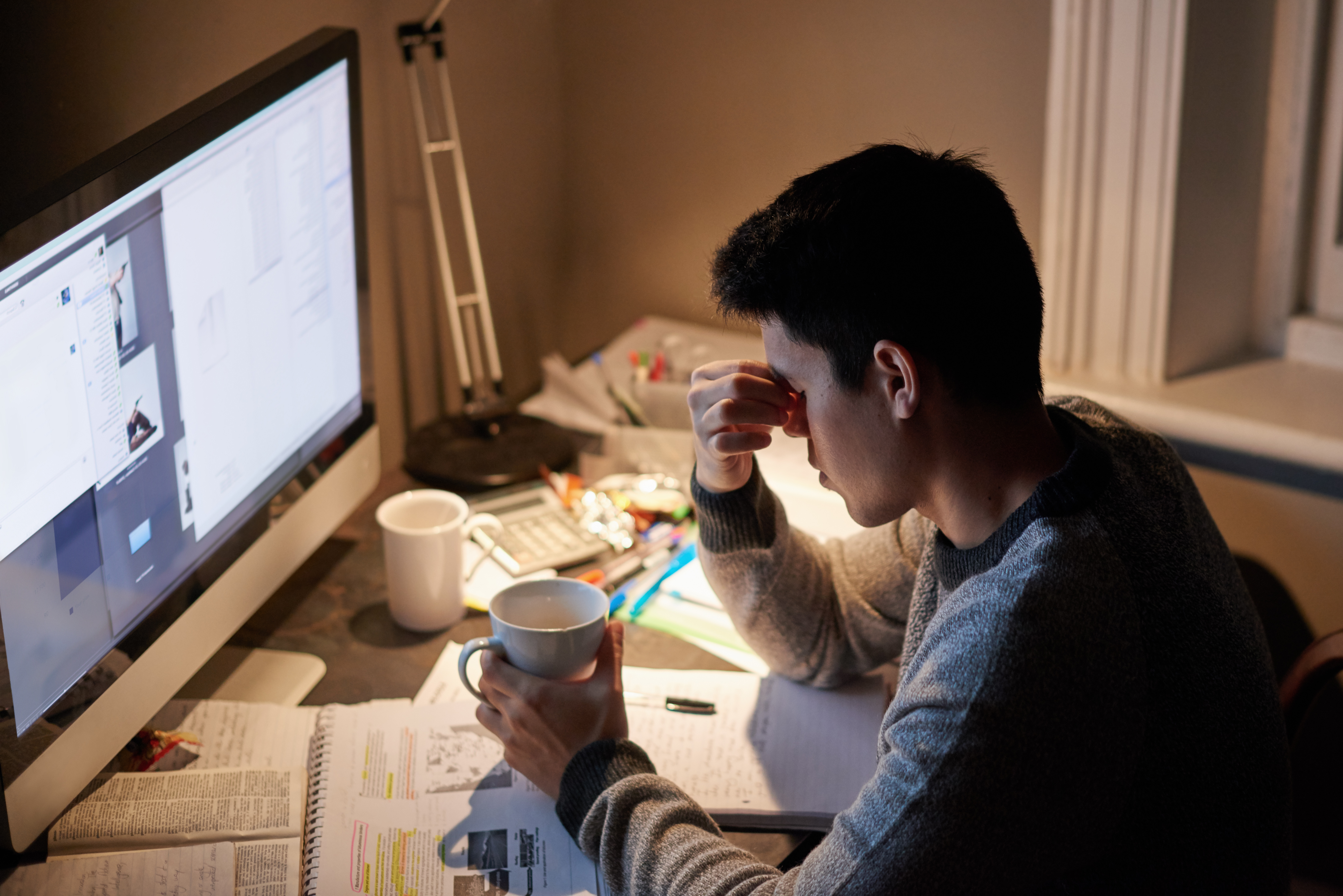 A young man sits at his desk in front of a desktop computer screen looking upset as he holds a mug