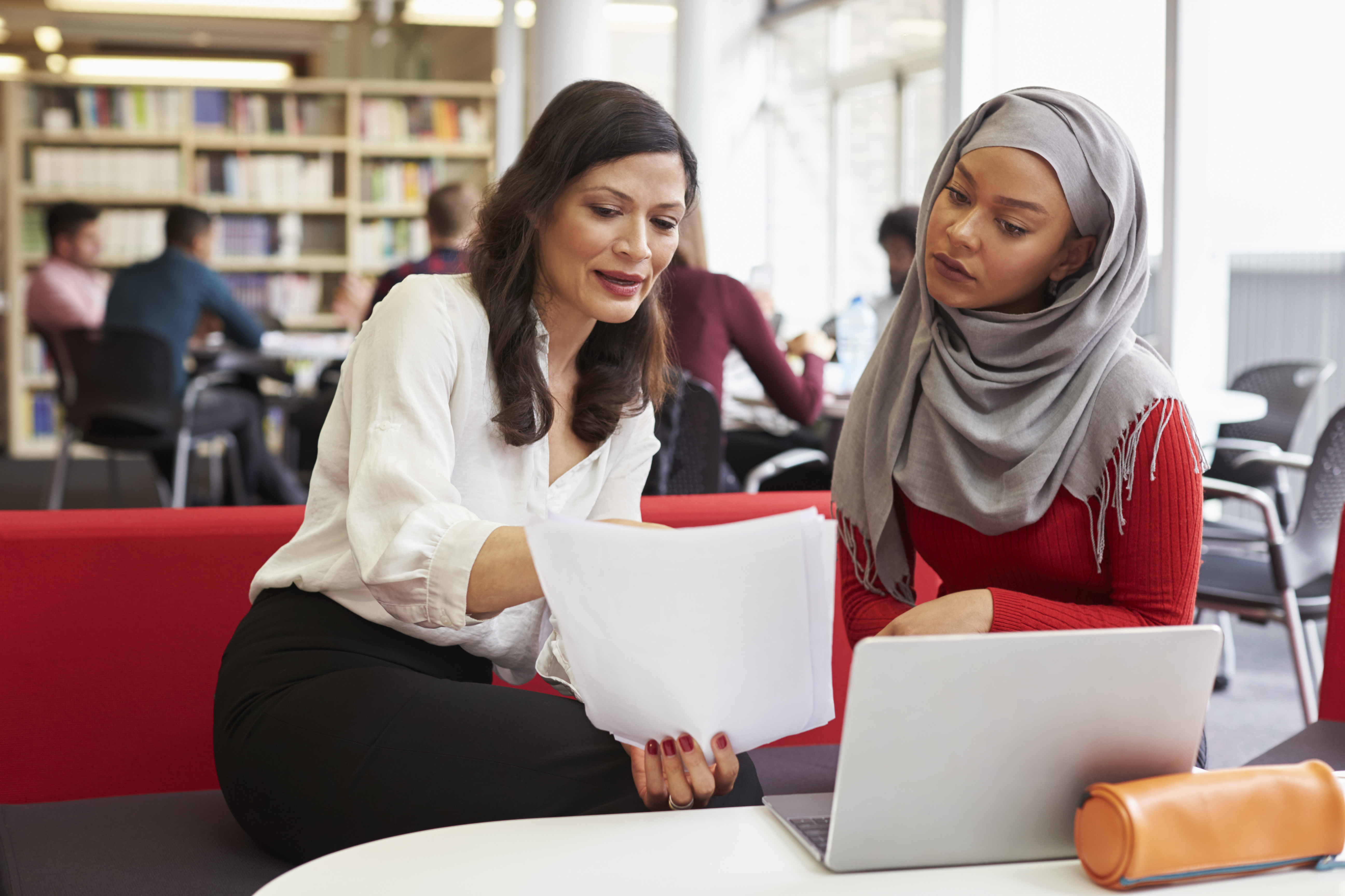 A woman sits next to another woman and they both look at pieces of paper being held in front of them