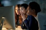 Three women sit together staring at a desktop computer screen and looked shocked