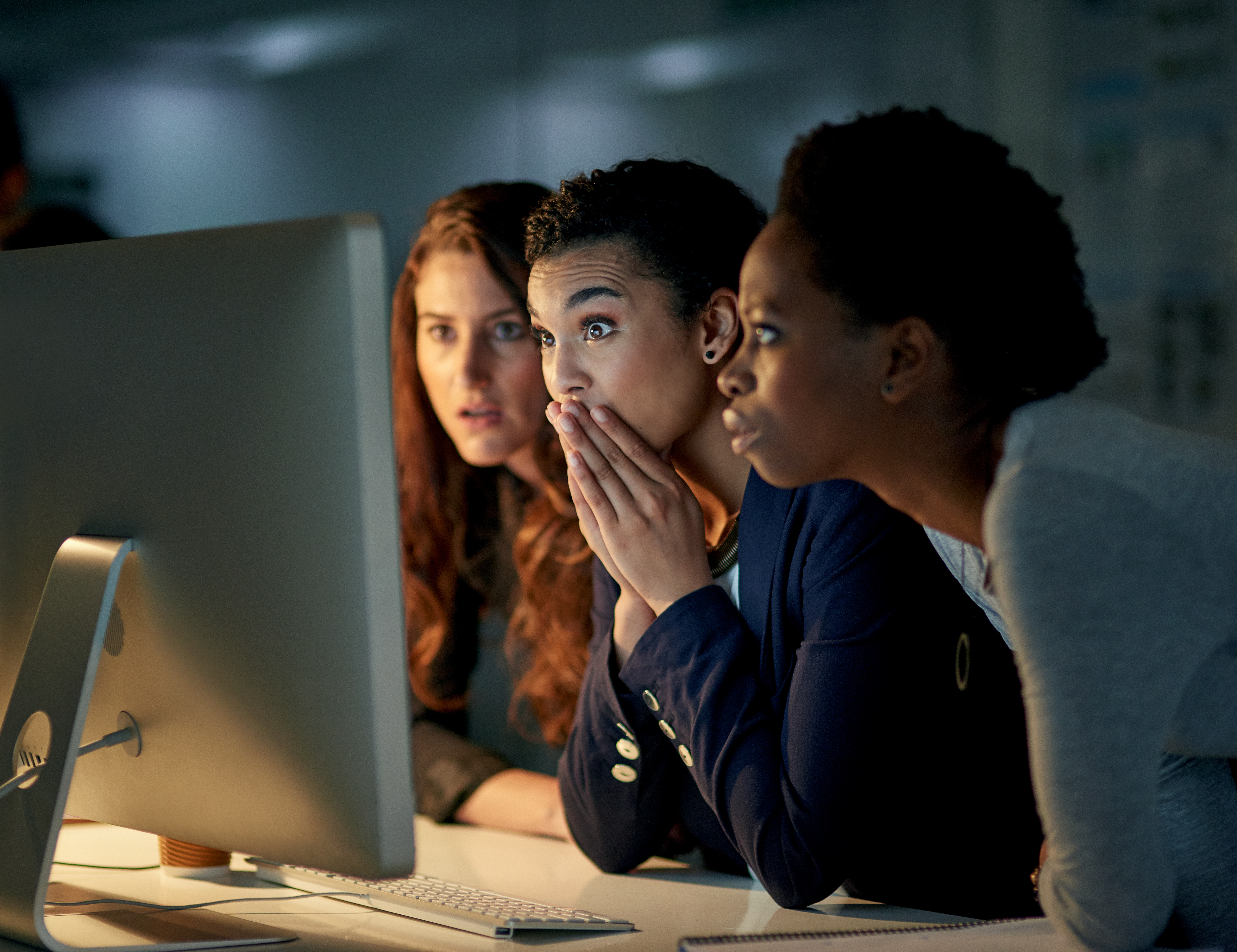 Three women stare at a desktop computer screen in a dark room and look shocked