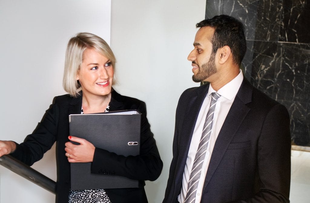 A man in a suit stands next to a woman in a smart black blazer who is holding a folder