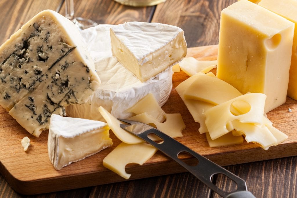 A selection of cheeses, including brie, cheddar and blue stilton, are arranged on a wooden chopping board on a wooden surface