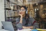 Young woman sitting at a desk staring out of the window with a laptop and books in front of her
