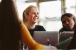 A girl sits at her laptop looking happy as she smiles and looks over the top of the laptop at something else