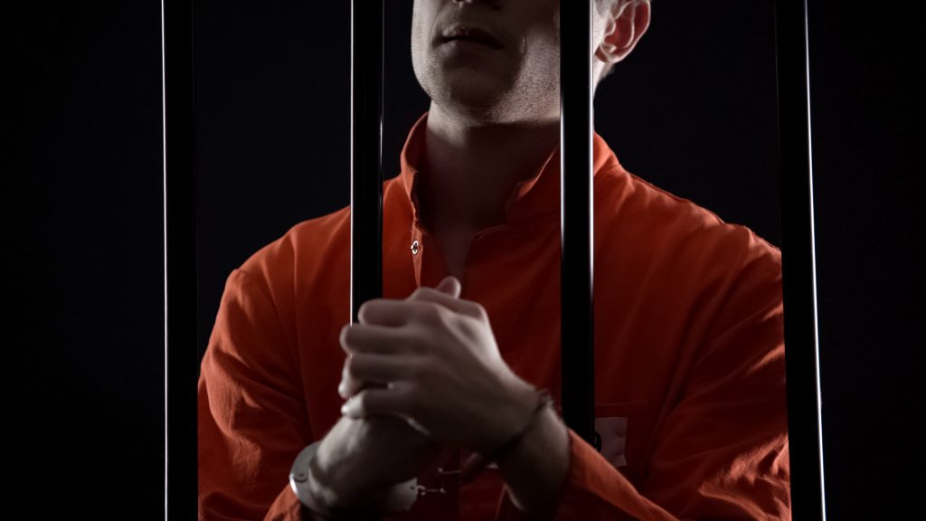 Criminal in orange jumpsuit and handcuffs behind bars