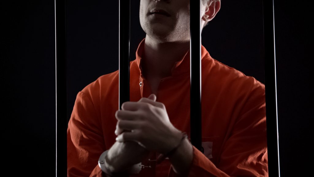 Criminal in orange jumpsuit and handcuffs behind bars