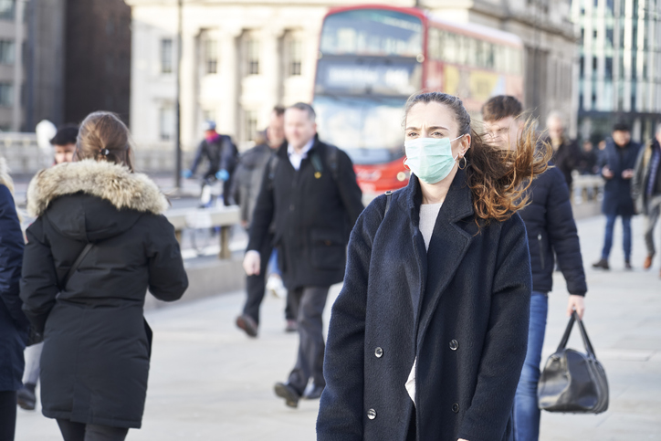 Woman wearing a facemask walking down a london street with a red bus and other pedestrians behind her