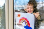 Little boy sticking a picture of a rainbow on his window during the COVID-19 pandemic