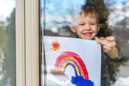 Toddler sticking a painting of a rainbow on his window during the COVID-19 pandemic