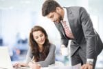 A woman is sat at her desk with a laptop open and a man stands next to her looking at the work on the desk
