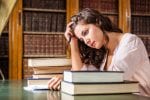 A girl sits at a table surrounded by books and looks sad about all the work she has to do