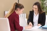 A woman sits at a desk with another woman and they look at a document on the table in front of them