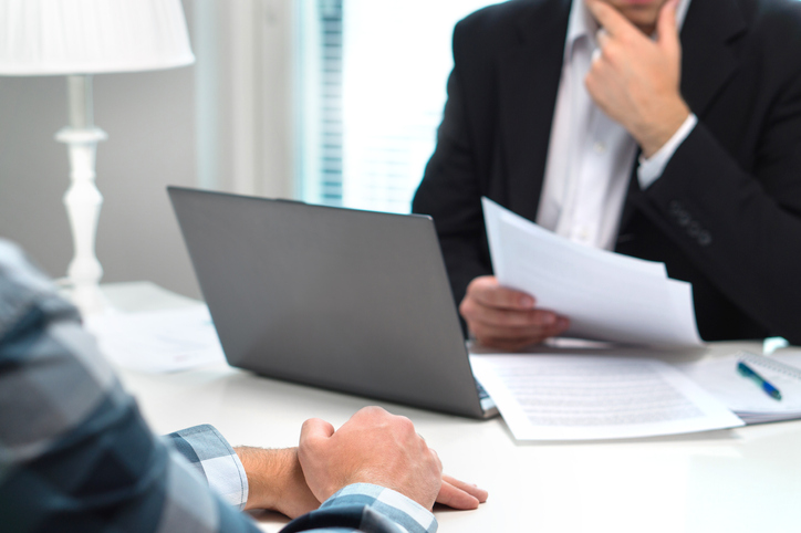 A person in a suit sits at a table with their laptop and paper surrounding them as they interview another person for a job