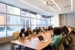 A group of workers sit around a meeting table in an office with open laptops in front of them