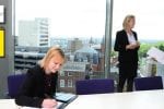 Two lawyers standing in front of a window discussing something, another is sat at the table signing a document